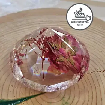Ein gegossenes Objekt in Form eines Diamanten, welcher eine rote Blume beinhaltet und auf einer Holzscheibe liegt